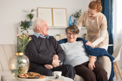 cheerful senior couple sitting on a couch with a tender caregiver standing next to them and putting a blue blanket over the woman's shoulder in a bright living room